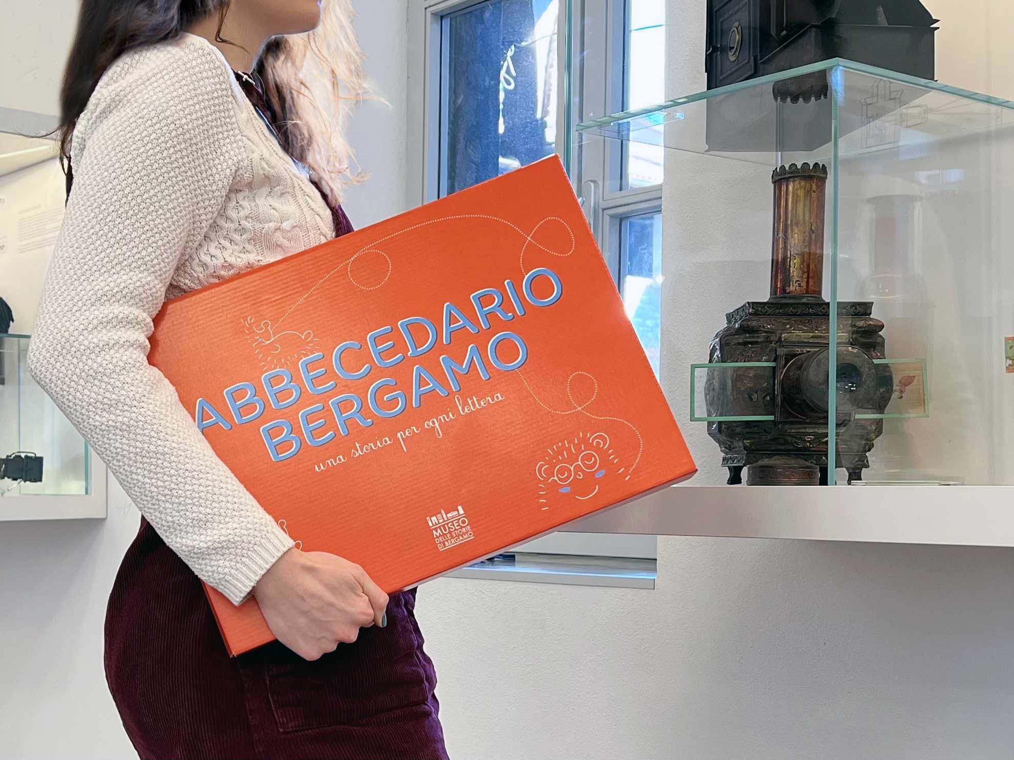 una ragazza tiene in mano una valigetta rossa con scritto "Abbecedario Bergamo"