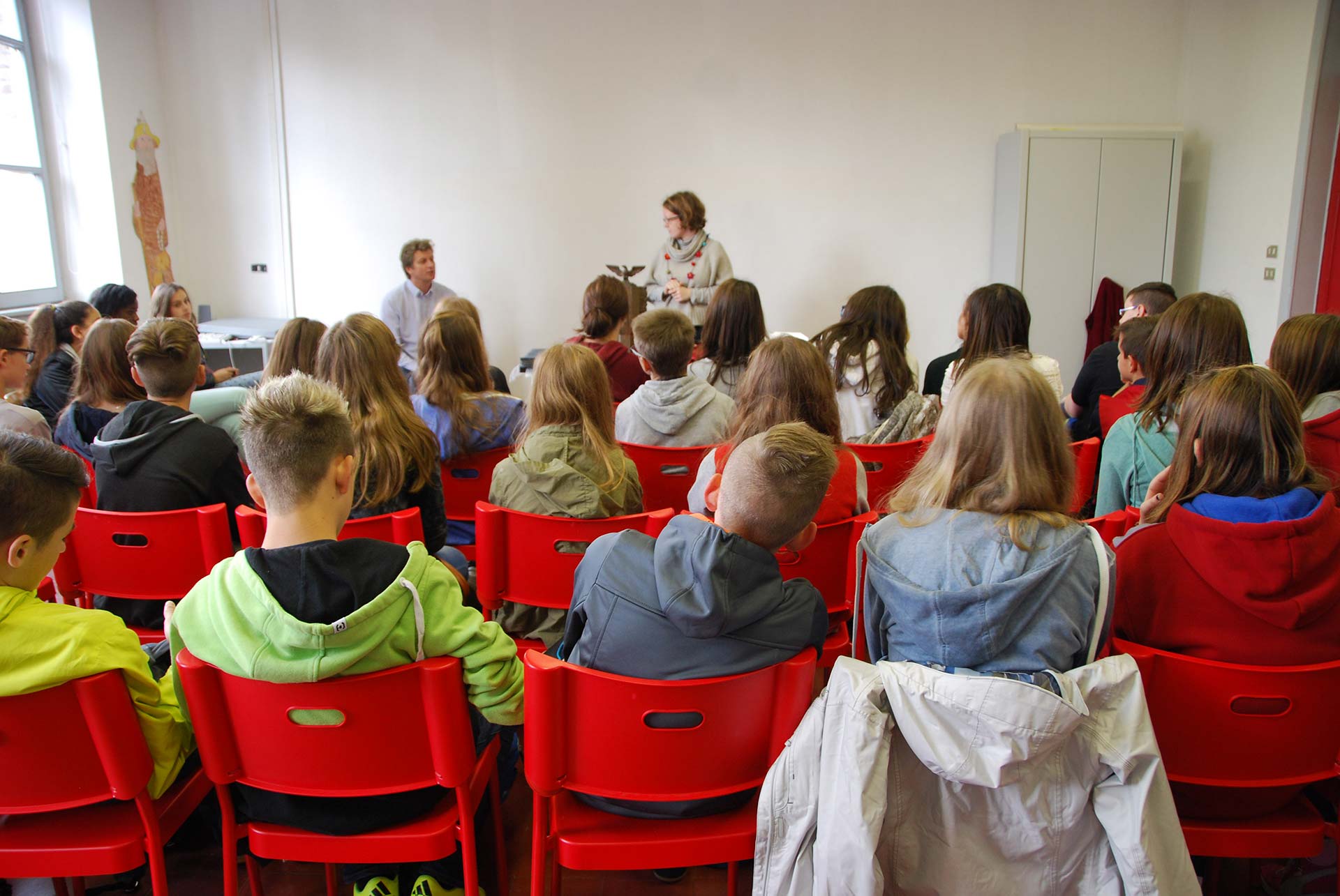Studenti di spalle seduti sopra delle sedie rosse ascoltano due persone che raccontano